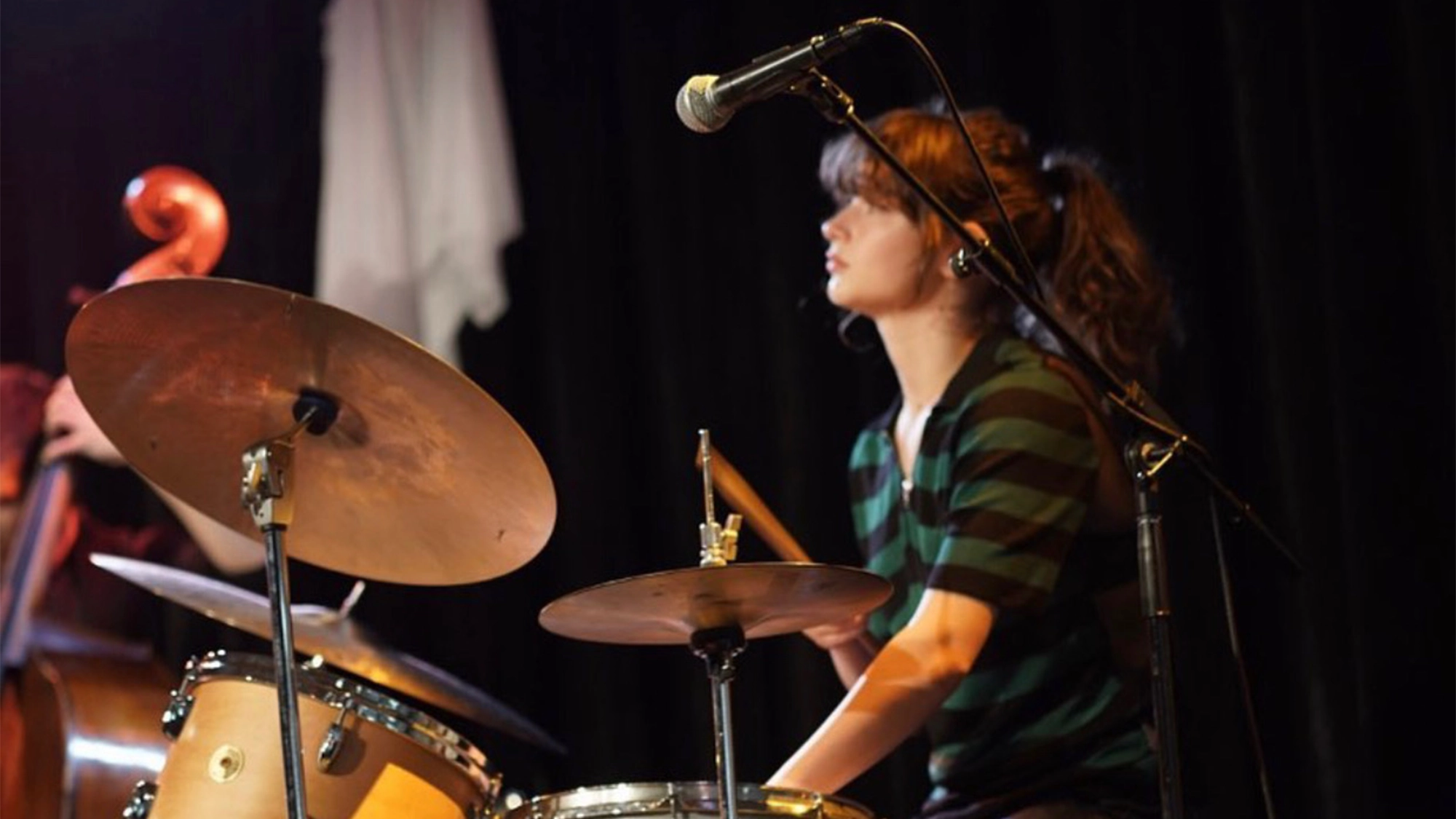 Ruby Laks site image 25 A young woman in a green and black striped shirt plays a drum set on stage, holding drumsticks. A microphone is positioned above her, and a double bass is partly visible on the left. The background is dark, suggesting a live music performance.