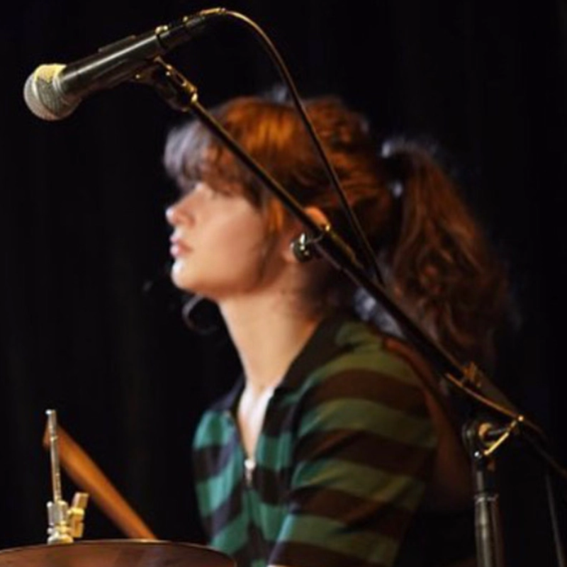 A young woman with wavy brown hair in a ponytail sits at a drum set, wearing a green and black striped shirt. She appears to be playing drums, with drumsticks in hand. A microphone is in the foreground, and the background is dark and out of focus.