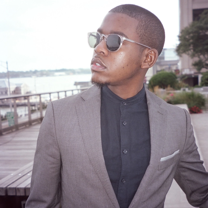 A man wearing sunglasses and a gray suit with a black shirt stands outdoors by a waterfront. He looks to the side with a serious expression. The background features railings, greenery, buildings, and water under a cloudy sky.