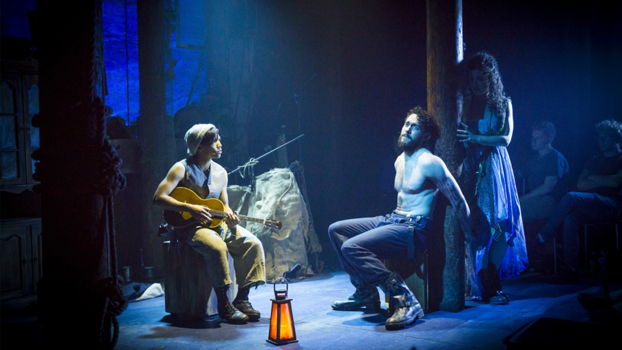 SeaWife site image 25 On a dimly lit stage at the 10th Anniversary Concert of SeaWife, a shirtless man is tied to a post, slumped with head turned. The Lobbyists' musician sits nearby playing guitar as a woman stands close, partially in shadow. A lantern glows on the floor.