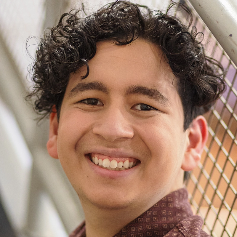 Sergio Guerra, a young person with curly dark hair, smiles warmly at the camera. They are wearing a patterned brown shirt and stand in front of a metal fence, with the background softly blurred and bright, natural lighting.