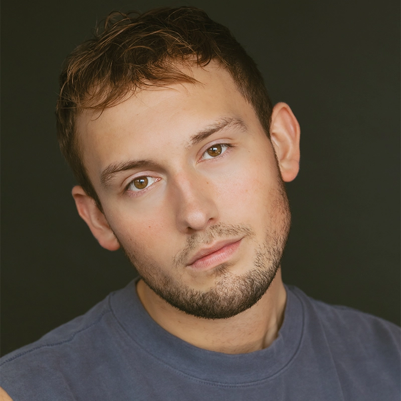 A young man, Seth Remington, with short brown hair and light facial stubble looks directly at the camera with a neutral expression. He is wearing a sleeveless, faded gray-blue shirt against a dark, plain background that draws focus to his face.