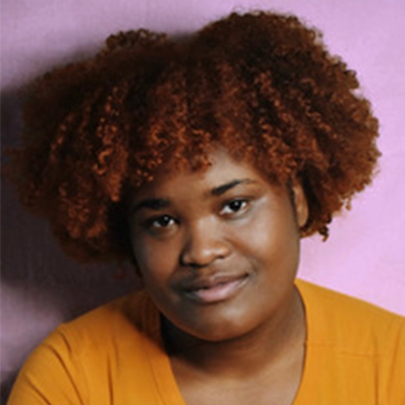 Marguerite A. Boone, a woman with brown, curly hair and medium-dark skin, smiles softly at the camera. She wears a mustard-yellow top and sits before a light purple background, with soft lighting accentuating her face and hair texture.