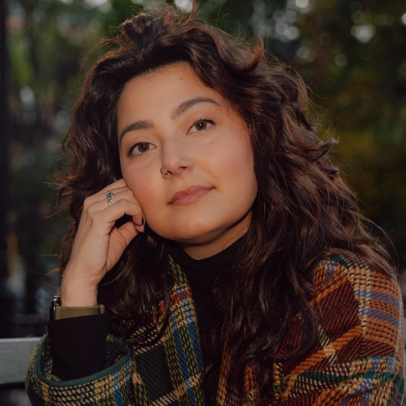 Amanda Harris, with wavy brown hair and a nose ring, rests her chin on her hand while gazing at the camera. She wears a plaid coat in earthy tones over a black turtleneck, set against a backdrop of blurred outdoor greenery.