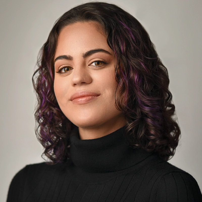 Camelia Elías Sierra, a young woman with shoulder-length curly brown hair and purple highlights, smiles slightly. She wears a black turtleneck sweater and faces the camera against a plain, light gray background with soft, even lighting.