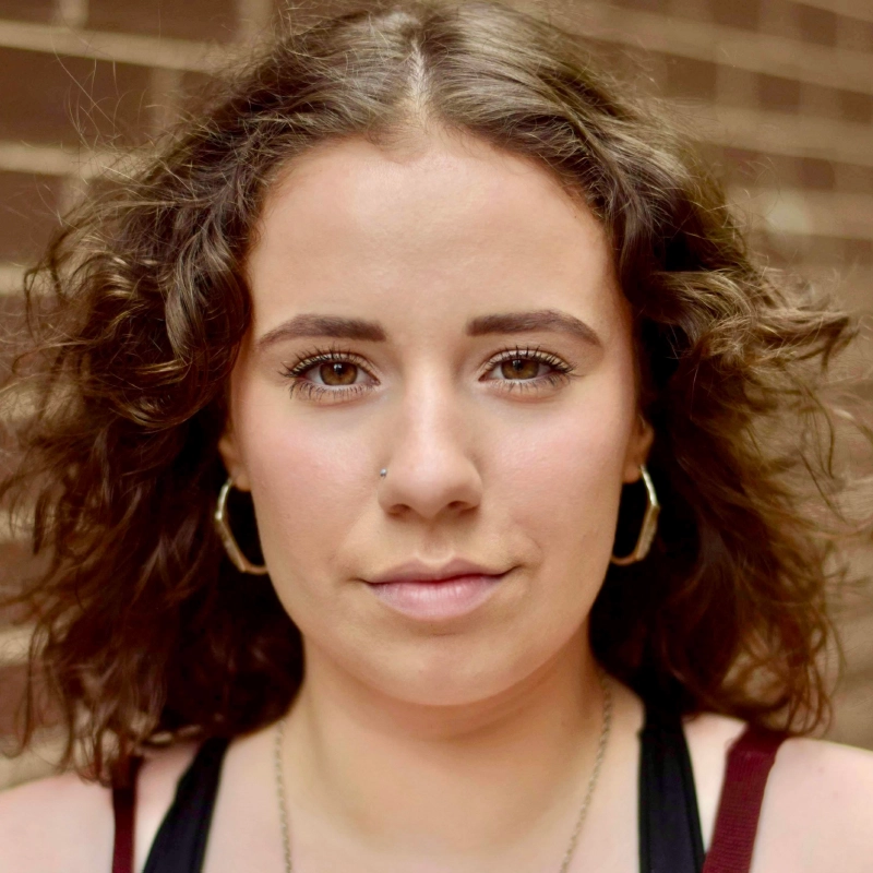 A young woman, Olivia Luzquinos, with medium-length curly brown hair stands in front of a blurred brick wall. She has a neutral expression, wears hoop earrings, a nose stud, subtle makeup, and a dark tank top with thin straps.