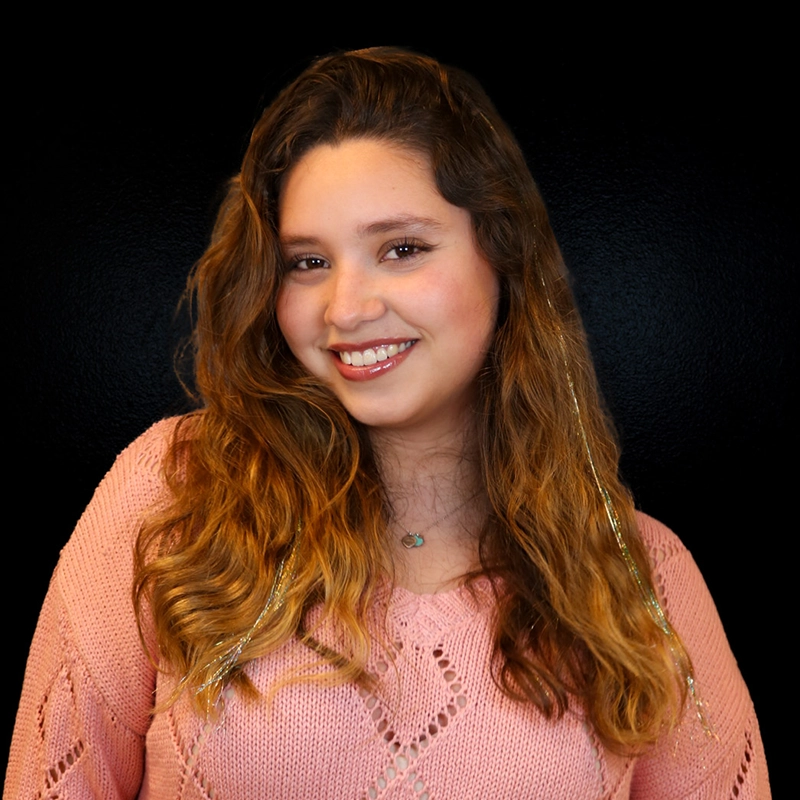 A young woman with long wavy brown hair, wearing a pink knit sweater and turquoise necklace, smiles at the camera. She has shiny tinsel strands in her hair. The background is solid black. This could be Jaeden Riley Juarez.