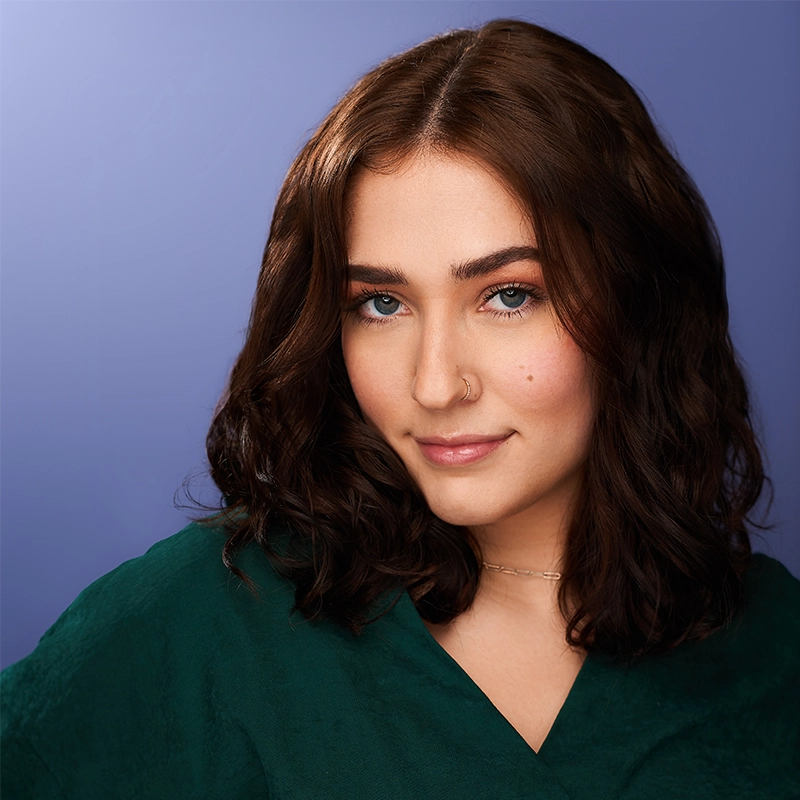 Emma Wallace, a young woman with wavy brown hair and light blue eyes, smiles softly. She has a nose ring, subtle makeup, and wears a green top with a delicate necklace. A smooth purple gradient background enhances her calm, confident expression.