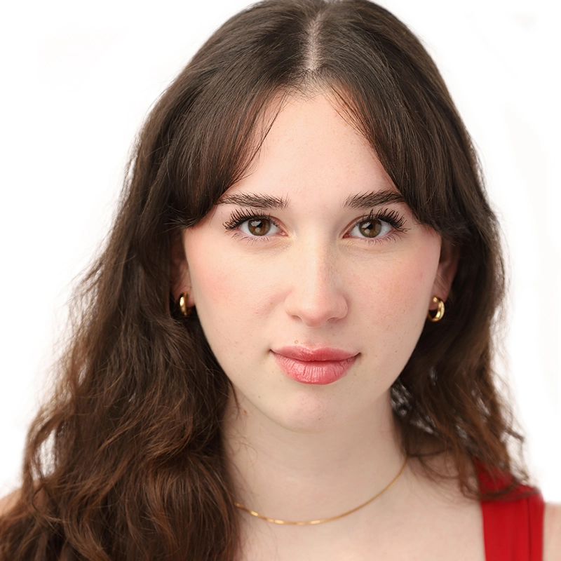 A young woman with long brown hair and fair skin gazes directly at the camera. Marielle Utayde wears gold hoop earrings, a thin gold necklace, and a red top. The plain white background highlights her natural makeup and calm expression.