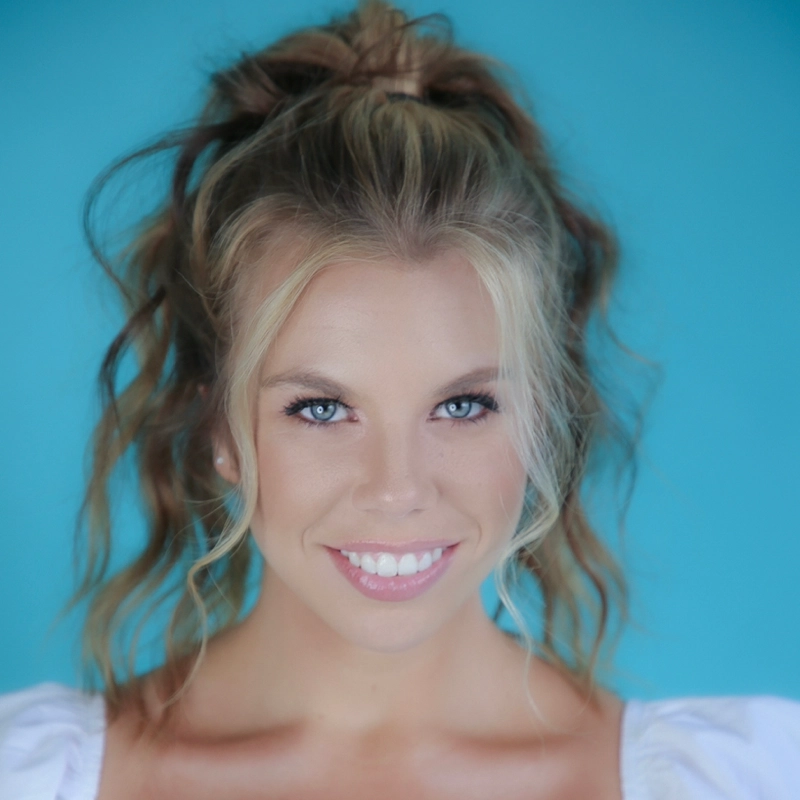 A young woman with wavy blonde hair in a high ponytail smiles at the camera. She has blue eyes, light skin, and wears a white top. The background is a solid turquoise blue, creating a vibrant and cheerful portrait.