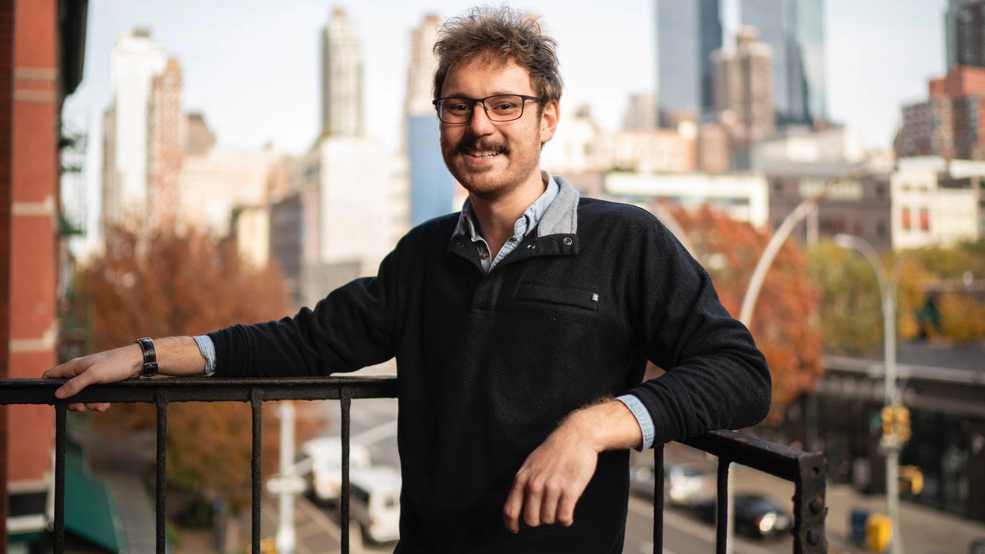 The Music of Eddie Bean - Show Art A man with glasses, a mustache, and curly hair—resembling Eddie Bean—stands outdoors, leaning on a black metal railing. He wears a black sweater over a collared shirt. City buildings, autumn trees, and music from the busy street fill the brightly lit scene.