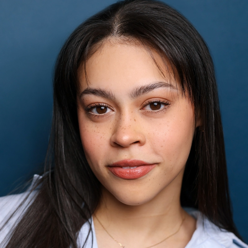 Lizzy Tucker, a young woman with straight dark hair and light brown skin, looks into the camera with a slight smile. She has brown eyes, natural makeup, and a few freckles, wearing a pale blue shirt and gold necklace against a dark blue background.