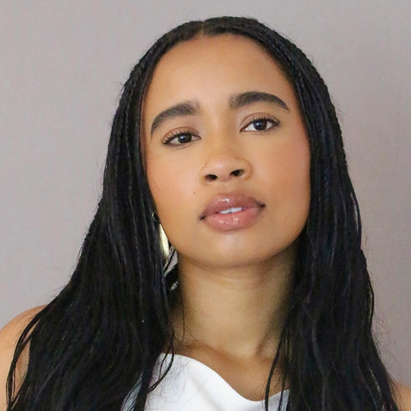 Marielle Utayde, a young woman with long, straight black hair in braids, poses against a plain light gray background. She wears a sleeveless white top, small gold hoop earrings, and has natural makeup with glossy lips and defined eyebrows.
