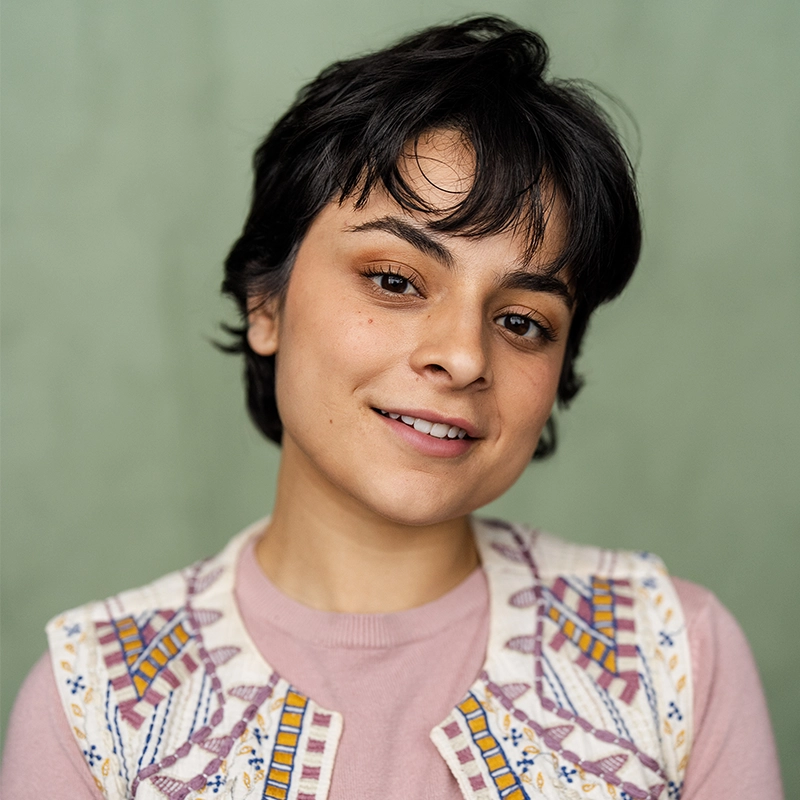 A young person with short dark hair smiles softly. They wear a pale pink shirt and a cream-colored embroidered vest. The plain, muted green background and soft lighting highlight Valeria Aceves's natural features and warm complexion.