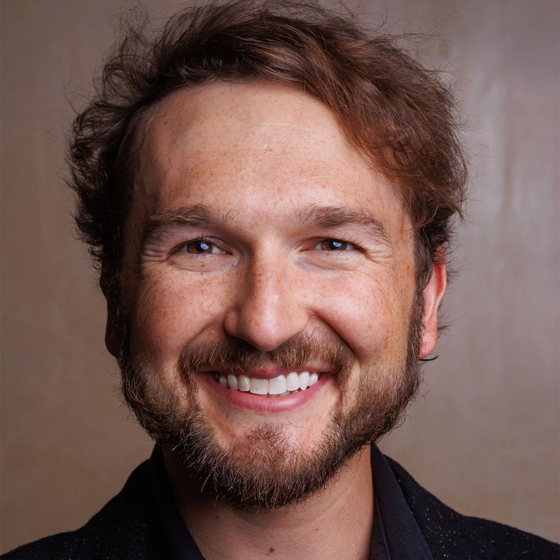 A man with wavy brown hair and a full beard smiles warmly at the camera. He has light skin, brown eyes, and is wearing a dark shirt and jacket. The background is plain and brown, keeping the focus on his friendly expression.