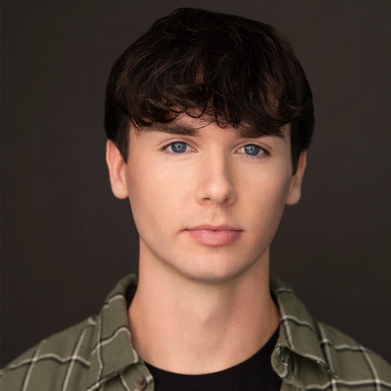 Josiah Jennings, a young man with fair skin, blue eyes, and short wavy brown hair, looks directly at the camera. He wears a green plaid shirt over a black t-shirt. The dark, plain background highlights his face.