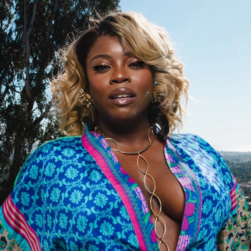 A confident woman with curly, shoulder-length blonde hair poses outdoors. She wears a blue and pink patterned robe with a deep neckline and a gold chain necklace. Trees and a clear blue sky are in the background.