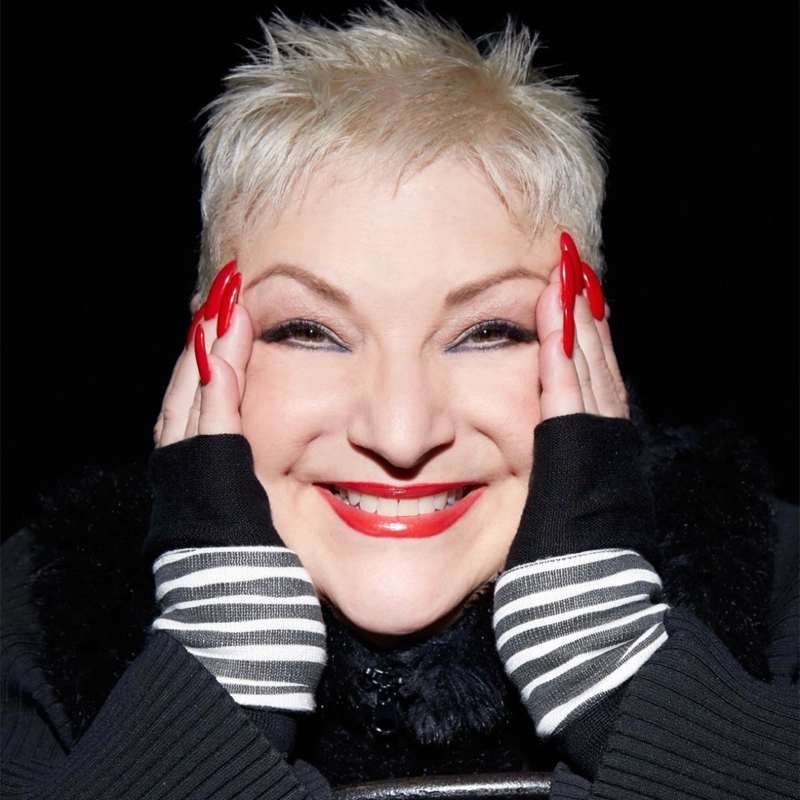 A smiling woman with short, spiky platinum-blonde hair holds her face with both hands, showing long red nails. She wears black fingerless gloves with white stripes and a fluffy black scarf. Her makeup includes bold red lipstick and winged eyeliner. The background is black.