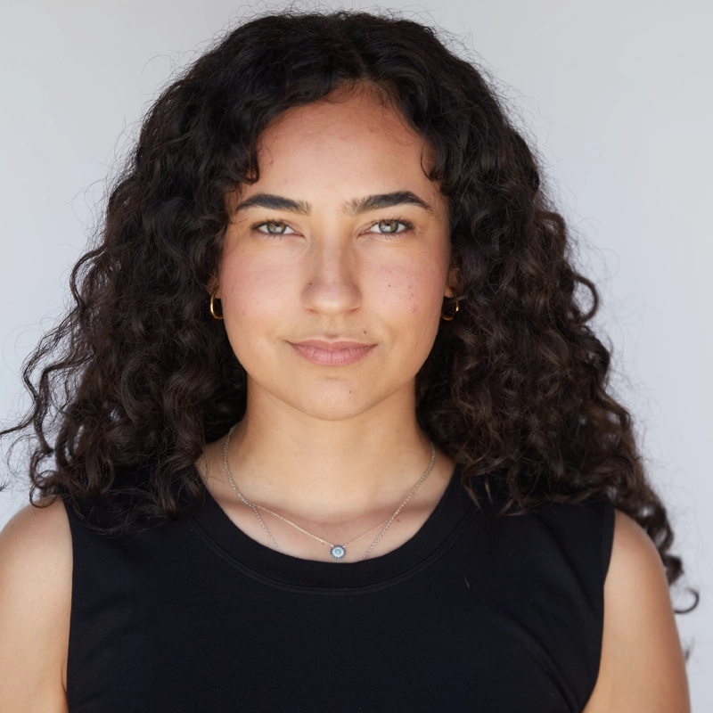 Alexa de la Pena, with medium skin, long curly dark hair, and hazel eyes, faces the camera. She wears a sleeveless black top, gold hoop earrings, and a silver necklace with a small blue pendant against a plain light background.