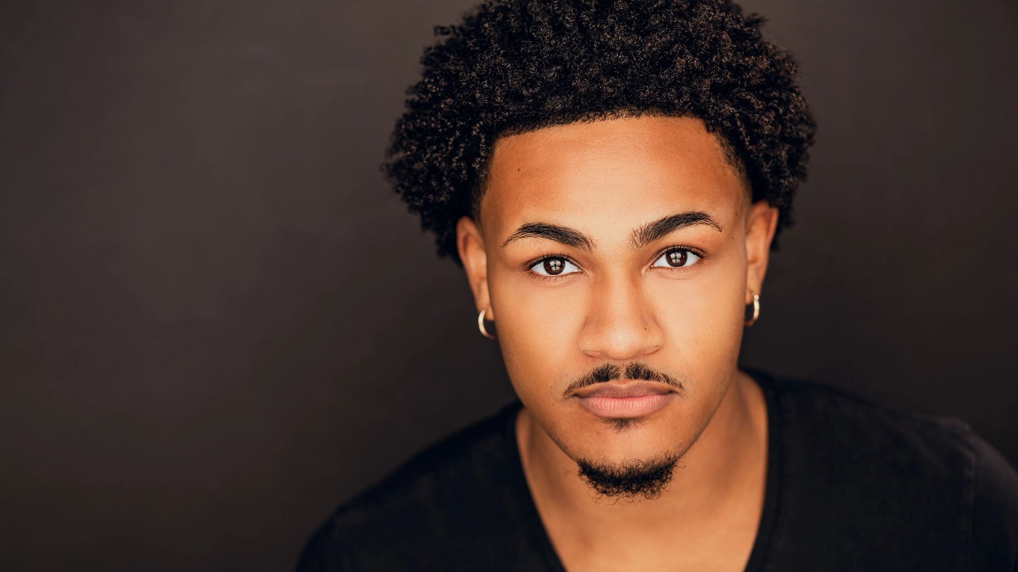 A young man with short curly dark hair and a trimmed goatee gazes confidently at the camera. Aaron Patterson wears small hoop earrings and a black shirt. The dark brown background puts focus on his expressive eyes and calm expression.