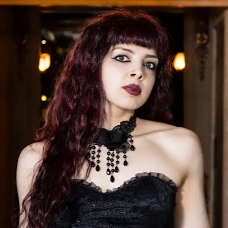 A woman with long, wavy dark red hair and straight bangs stands indoors. She wears dramatic black eyeliner, dark lipstick, a strapless black lace dress, and a statement black beaded necklace. Warm lights and a blurred background are visible behind her.