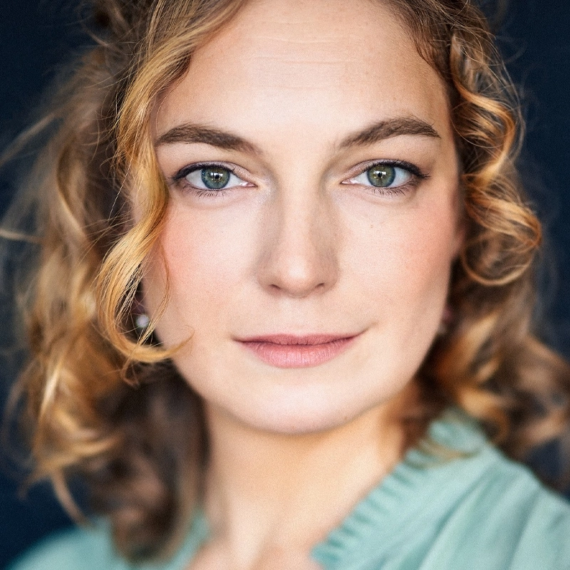 Gloria Bangiola, a woman with wavy blonde hair and green eyes, looks directly at the camera with a calm expression. She wears a pale green top with a ruffled neckline. The dark background makes her face stand out in the soft lighting.