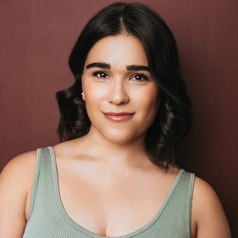 Jenna Barricklo, a young woman with medium-length dark wavy hair and smooth skin, smiles softly in front of a burgundy background. She wears a sage green tank top and small stud earrings, with natural makeup and defined brows.