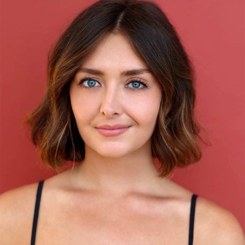 Kailey Boyle, with short, wavy brown hair and blue eyes, stands against a solid red background. She wears a black spaghetti strap top and smiles softly at the camera, giving a warm, approachable expression.
