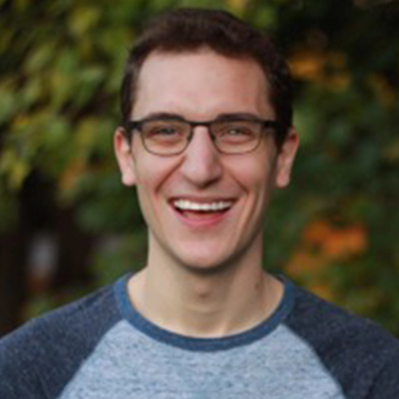 Brendan Stackhouse, a young man with short brown hair and glasses, smiles broadly. He wears a blue and gray raglan shirt. The background is blurred, with green foliage and hints of autumn colors, suggesting an outdoor setting.