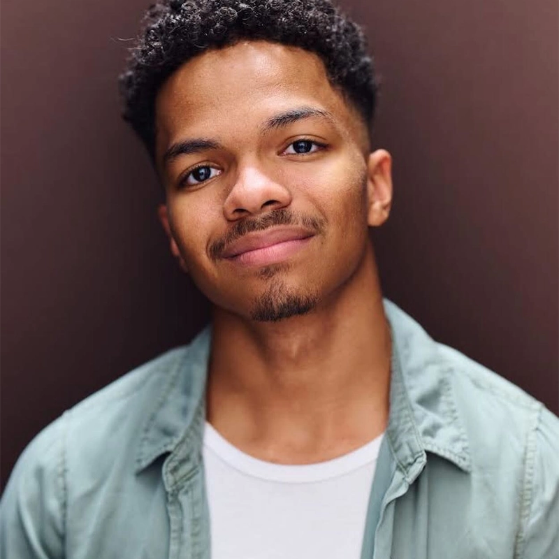 A young man with short curly hair and a trimmed goatee smiles gently at the camera. He wears a light green open shirt over a white t-shirt, standing against a plain dark brown background. The lighting is soft and even.