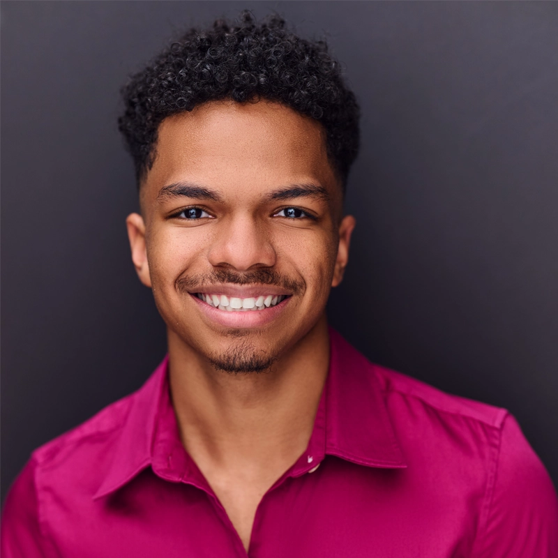 A young man with short, curly black hair and a trimmed goatee smiles at the camera. He wears a bright magenta button-up shirt and stands against a plain, dark background. The portrait has even lighting and a friendly, confident mood.