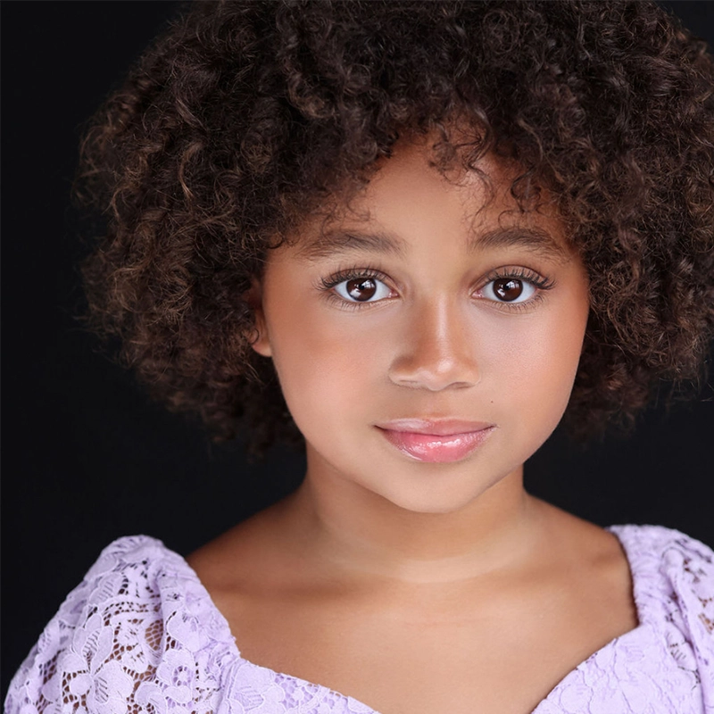A young child with curly brown hair and warm brown skin gazes softly at the camera. Wearing a light purple lace dress, they captivate with expressive eyes—a touching portrait inspired by Camilla Lockhart's evocative style.
