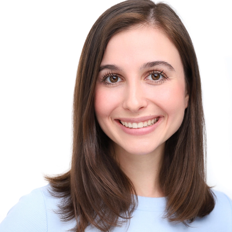 A young woman with straight, shoulder-length brown hair and brown eyes smiles at the camera. She is wearing a light blue top and is posed against a plain white background, creating a bright, professional headshot.