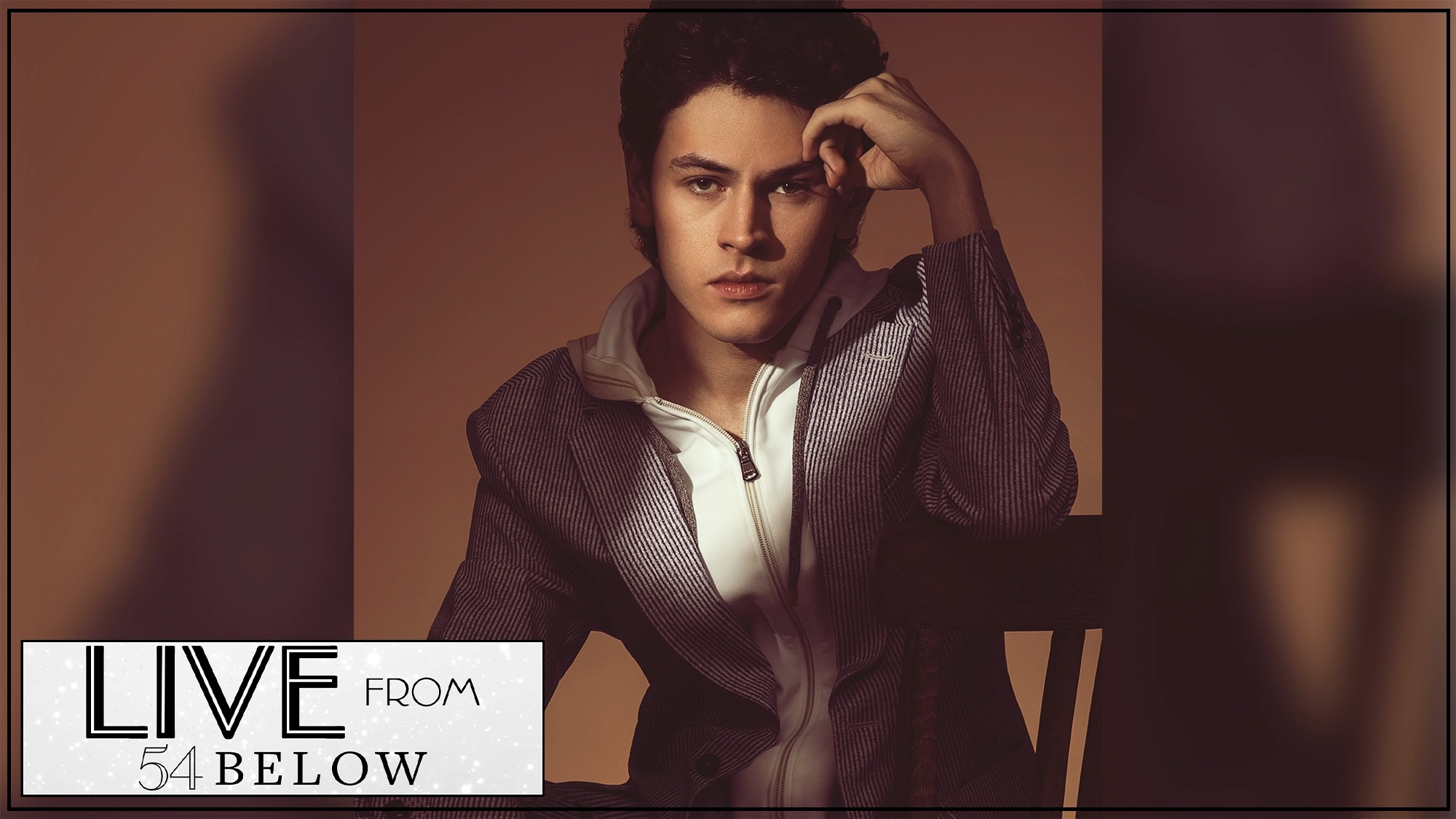 A young man with dark wavy hair sits on a chair, resting his head on one hand and gazing forward with a serious expression. He wears a striped jacket and white shirt. In the corner, “LIVE FROM 54 BELOW” promotes Casey Likes in a special livestream event.