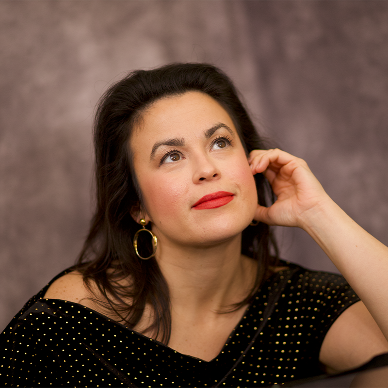 A jazz singer with dark brown hair and red lipstick gazes thoughtfully upward. She wears a black, off-the-shoulder top with gold dots and large gold earrings. Her hand rests against her face, the background softly blurred in gray and brown.