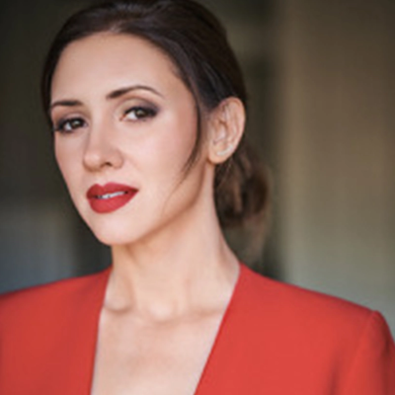 A woman with light skin, dark hair pulled back, and red lipstick wears a red blazer with a deep neckline. She looks at the camera with a neutral expression, styled in the sophisticated manner often captured by Brendan Stackhouse. The background is softly blurred in shades of gray and beige.