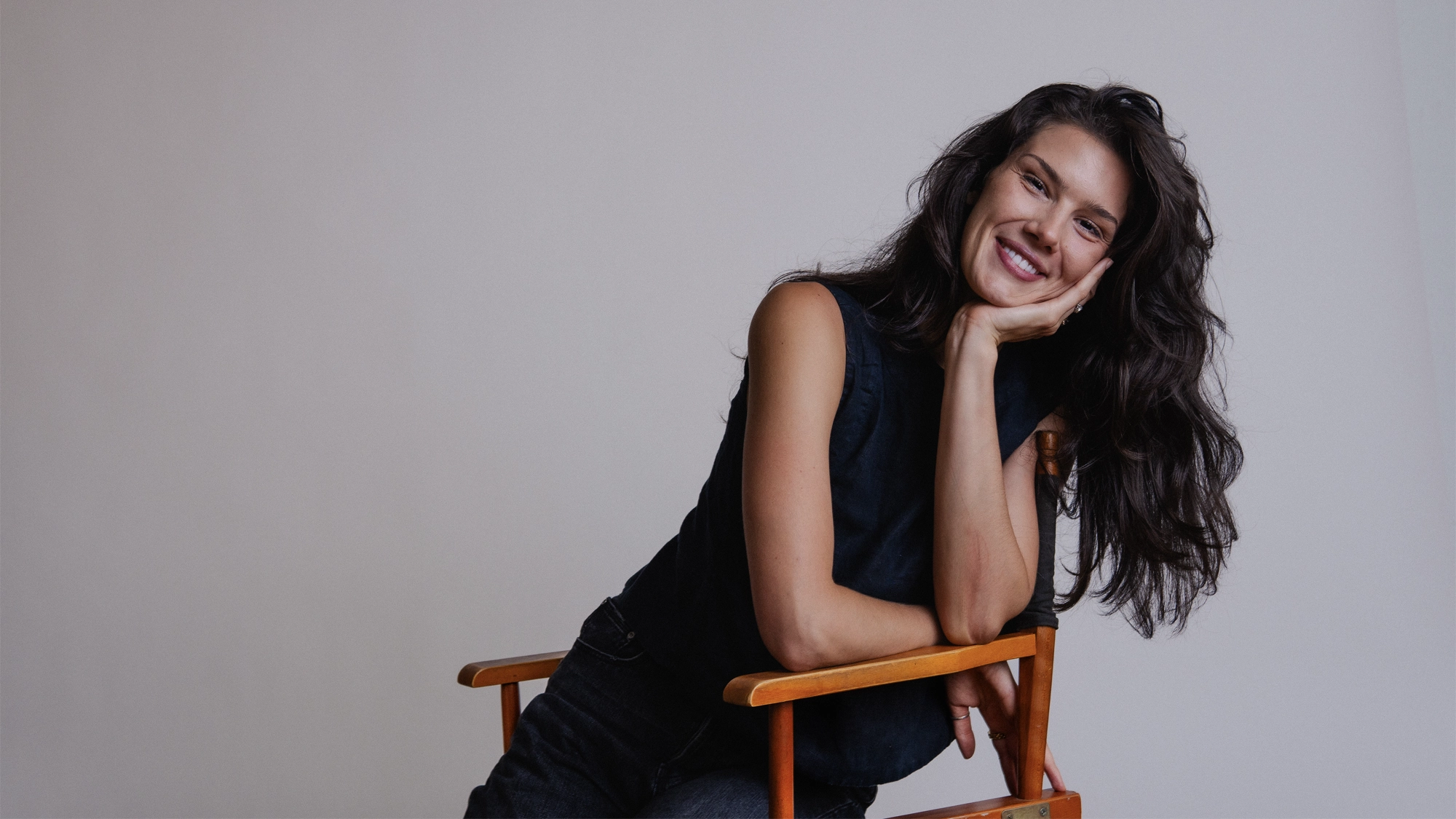 Christine Oczkus, a tall drink of me, sits sideways on a wooden chair, resting her chin on her hand and smiling warmly at the camera. She wears a sleeveless dark top and black jeans against a plain light-colored background.