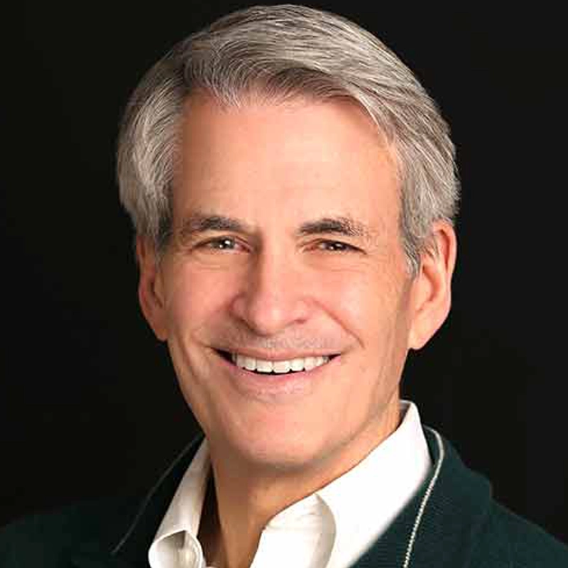 Michael Colby, a middle-aged man with short gray hair, smiles warmly at the camera. He wears a white collared shirt under a dark sweater, with a solid black background making his face stand out clearly.