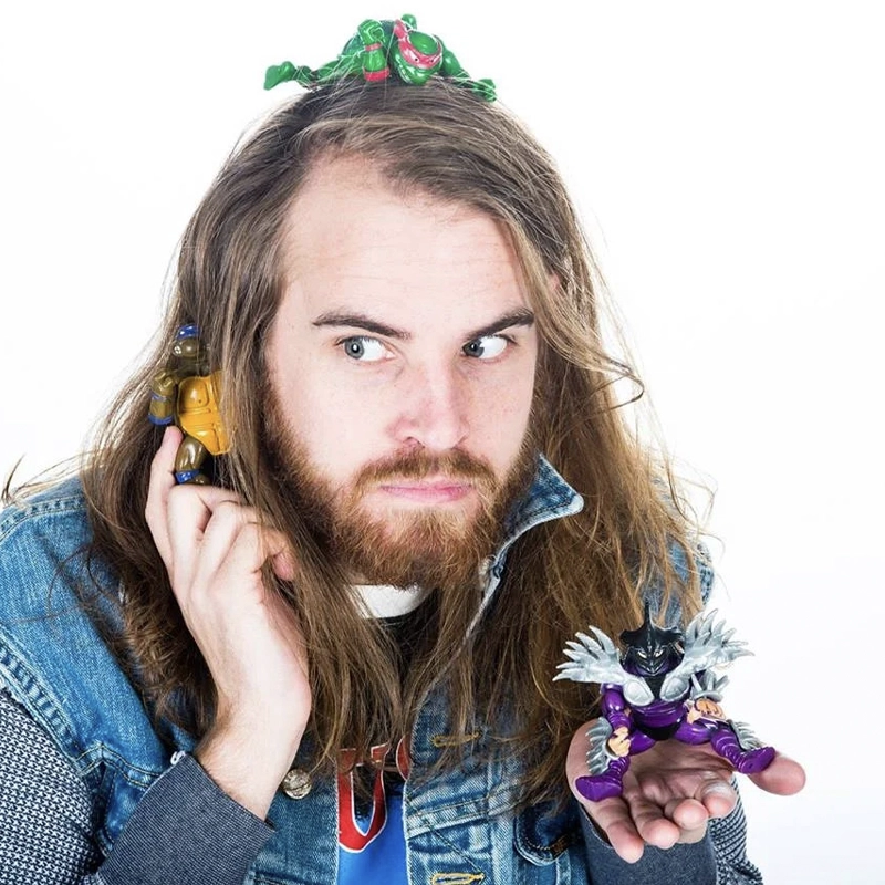A man with long brown hair and a beard, wearing a denim jacket, holds a Ninja Turtle toy to his ear and another villain toy in his palm. A green dragon toy rests on his head. He looks sideways with a curious expression against a white background.
