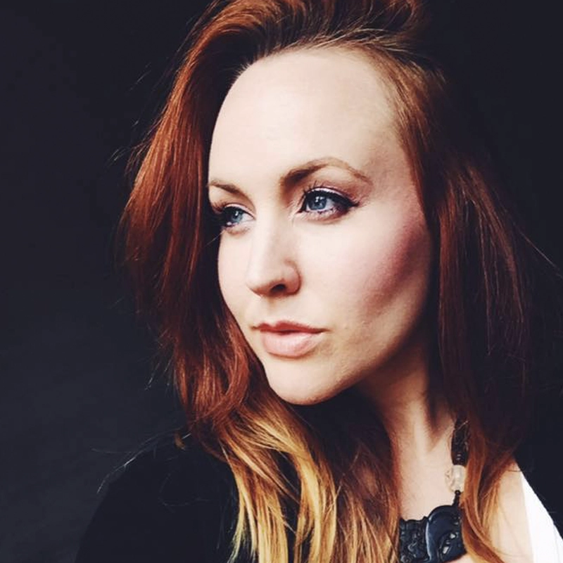 Dana Kelly Craig, a woman with long, wavy auburn hair and fair skin, looks thoughtfully to the side. She has blue eyes, subtle makeup, and wears a black top with a dark necklace. The dark background highlights her features and expression.