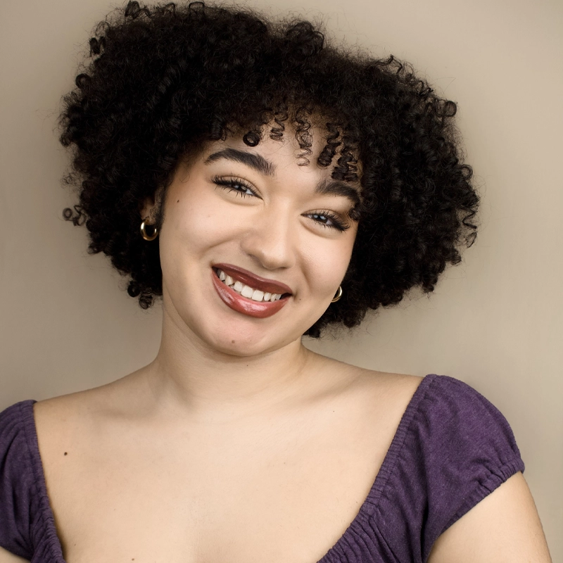 Maryana Crawley, a young woman with medium skin and curly dark hair, smiles at the camera. She wears gold hoop earrings, bold eyebrows, red lipstick, and a purple square-neck top against a soft beige background.