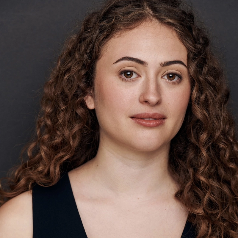 A young woman with long, curly brown hair and light skin looks calmly at the camera. She has full eyebrows, brown eyes, and neutral pink lips. She wears a dark sleeveless top and is posed against a plain dark background.