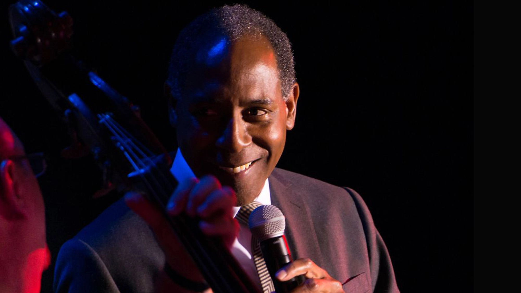 A man in a suit holds a microphone and smiles warmly at someone playing a double bass in the foreground. Dim red and blue stage lights highlight his face, creating an intimate atmosphere as they perform “Dedicated To You” on stage.