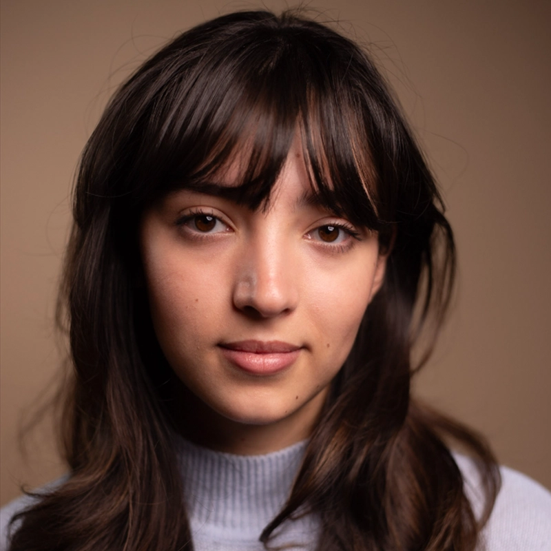 A young woman with long brown hair and bangs looks into the camera with a neutral expression. She wears a light gray turtleneck sweater. The background is a soft beige, and the lighting is warm and even.