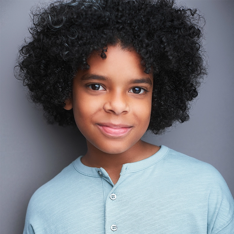 A young child with curly black hair and medium-brown skin smiles softly, wearing a light blue button-up shirt. The background is plain and gray, highlighting the child’s expressive eyes and gentle expression.