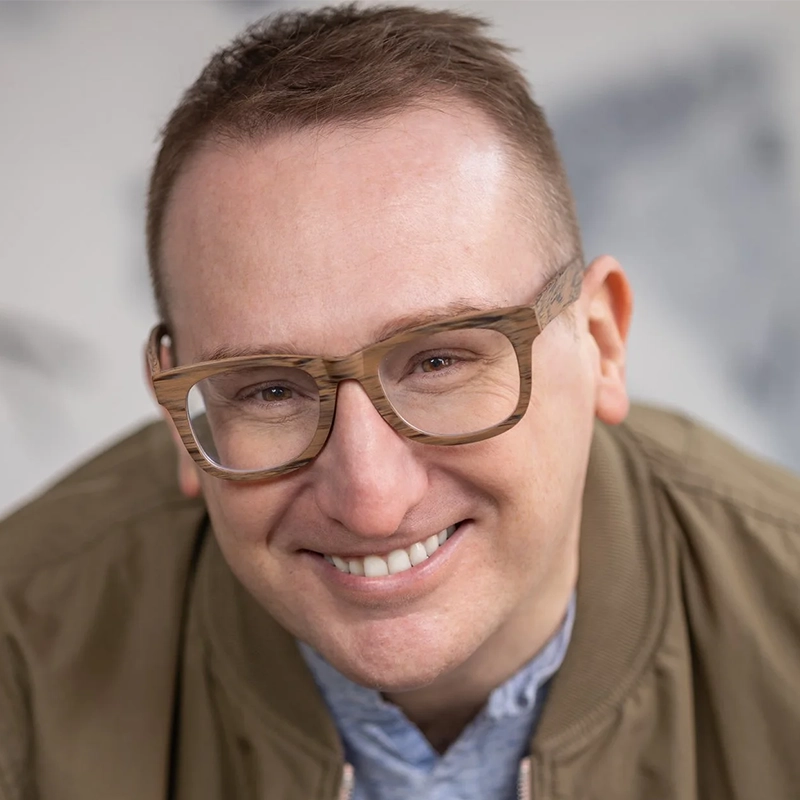 A man with short brown hair, wearing large, light-brown wooden glasses and a khaki jacket over a light blue shirt, smiles warmly at the camera. The background is softly blurred in gray and white tones, evoking a Shinnerie Jackson portrait style.