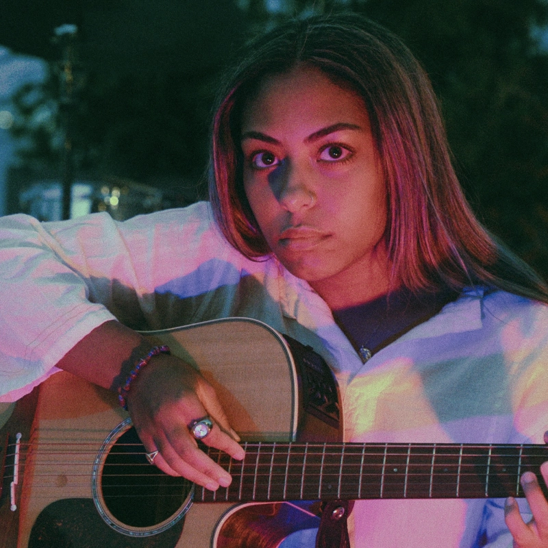 A young woman with long hair and an intense expression sits outdoors, illuminated by colorful pink and purple lighting. Daniella Faith wears a white shirt, bracelets, and a ring, holding an acoustic guitar with her fingers poised to play.