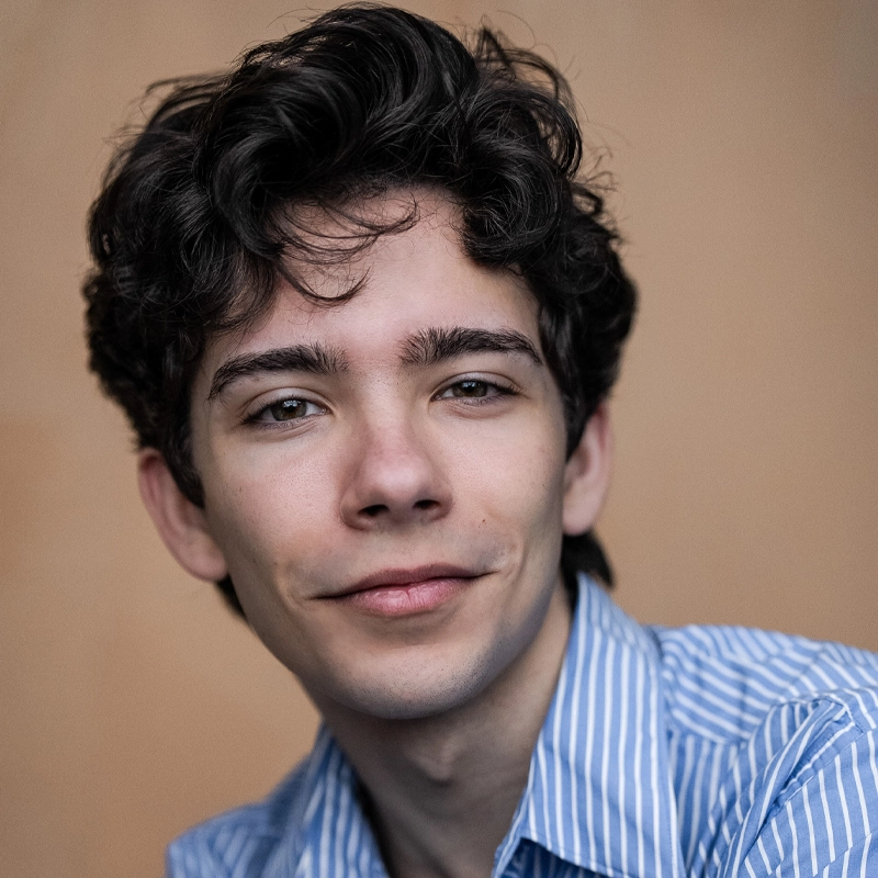 A young man with wavy dark hair, light skin, and brown eyes smiles subtly. Tommy Ferolano wears a blue-and-white striped collared shirt. The plain beige background keeps the focus on his relaxed, friendly expression.