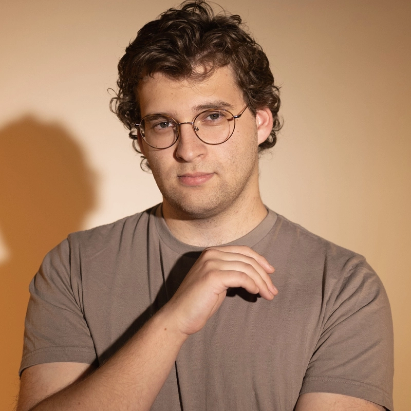 Zach Friedman, a young man with short, curly brown hair and glasses, stands against a beige background. Wearing a taupe t-shirt, he looks at the camera with a neutral expression, his right hand raised near his chest and his shadow cast on the wall.