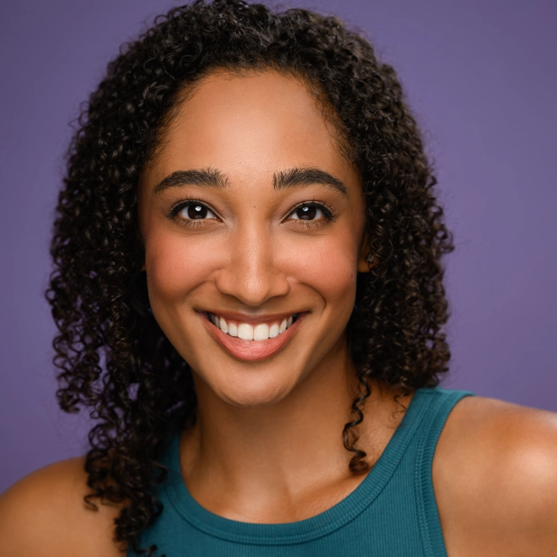 Elayna Garner, a young woman with medium brown skin and curly dark hair, smiles warmly. She wears a teal sleeveless top and is posed against a smooth purple background, looking directly at the camera under bright, even lighting.